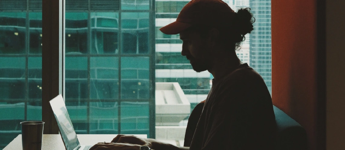 Silhouette of person typing on a laptop with office buildings in the back