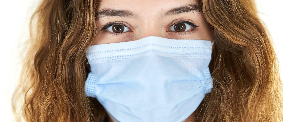 Caucasian woman with wavy hair wearing protective face mask looking at camera with white background