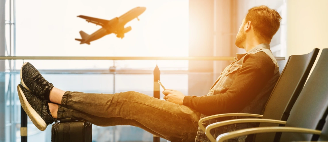 Man watching an airport take off while sitting in seat at the airport with legs resting on a suitcase 