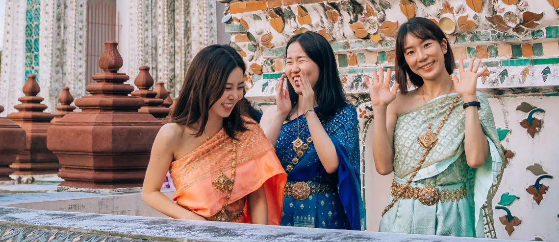 Three women in traditional Thai clothing smiling and waving at the camera