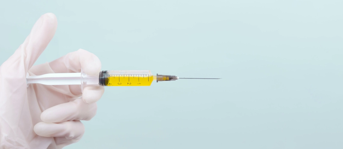 A doctor's hand in a white medical glove holds a syringe with a yellow medicine on a blue background